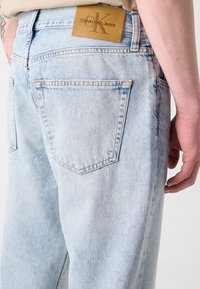 Svijetloplave traperice s klasičnim dizajnom pet džepova, izblijedjelom teksturom i istaknutom smeđom oznakom Calvin Klein Jeans na pojasu.