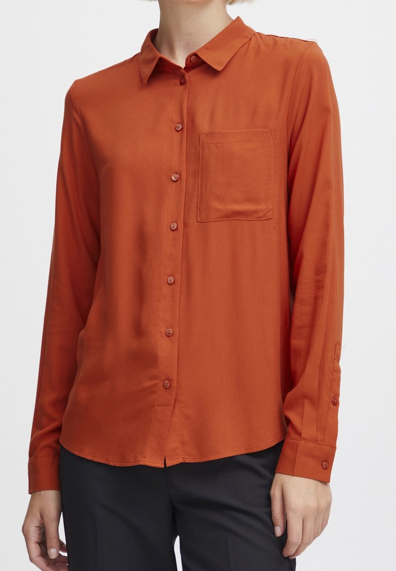 Chemise boutonnée couleur rouille en tissu doux, dotée d'une poche poitrine, de manches longues avec poignets à boutons et d'un col classique.