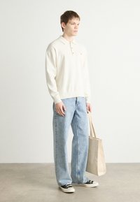 Ανοιχτό κρέμα πλεκτό polo shirt με γιακά, συνδυασμένο με ανοιχτό μπλε φαρδιά τζιν και μαύρα αθλητικά παπούτσια, κρατώντας μια μπεζ υφασμάτινη τσάντα.