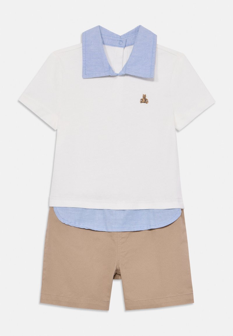 Polo blanca de manga corta con cuello azul, superpuesta a una camisa de tono azul claro, combinada con pantalones cortos beiges. Presenta un pequeño diseño bordado.