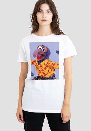 Frau trägt ein weißes T-Shirt mit einem bunten Motiv einer Puppe, die eine blaue Figur in einem gelben Hemd mit roten Chilischoten und karierten Hosen zeigt.