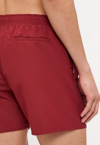 Shorts de bain bordeaux en tissu léger, avec une taille élastique et une poche arrière avec rabat. Texture douce, longueur au-dessus du genou.