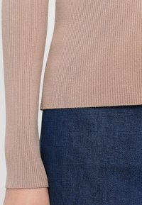 Pull beige côtelé à manches longues rentré dans un jean en denim bleu foncé, montrant les détails de la taille et des bras.