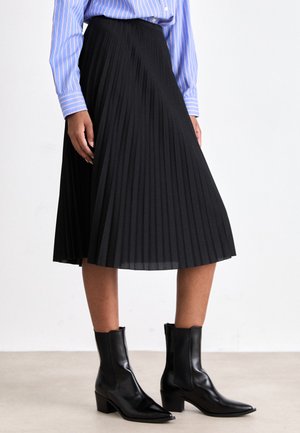 Jupe noire plissée mi-longue en tissu léger, associée à des bottines noires ajustées avec un bout pointu et un talon bloc.