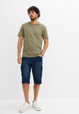Olivgrünes Baumwoll-T-Shirt kombiniert mit dunkelblauen Cargo-Shorts aus Denim, die mehrere Seitentaschen besitzen. Weiße Sneakers runden den Look ab.