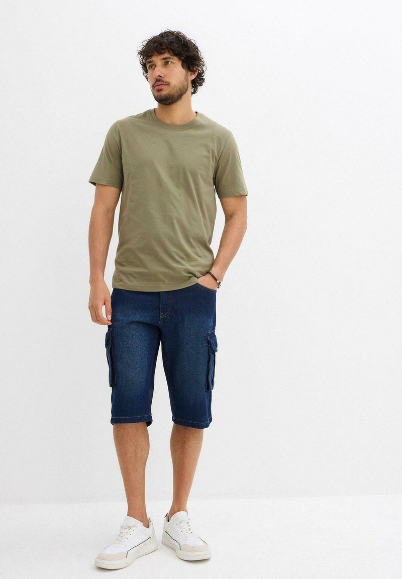 T-shirt in cotone verde oliva abbinata a pantaloni cargo in denim scuro con più tasche laterali. Sneakers bianche completano il look.