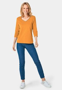 Senfgelbes V-Ausschnitt Langarmshirt kombiniert mit blauen Skinny Jeans und weißen Sneakers, vor einem weißen Hintergrund stehend.