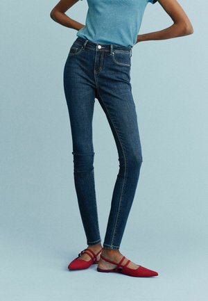 Jeans skinny en denim bleu foncé avec une texture lisse, associés à des ballerines rouges avec un design à lanières et un talon ouvert.