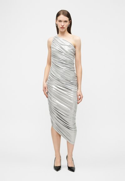 Γυναίκα που στέκεται με ασημί midi φόρεμα με έναν ώμο και πιέτες, και μαύρες μυτερές γόβες, μπροστά σε απλό φόντο.