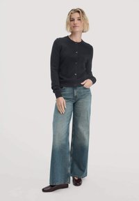 Schwarze Knopfkarte mit runder Ausschnitt, kombiniert mit hellblauen weiten Jeans. Das Model steht selbstbewusst mit den Händen in den Taschen.