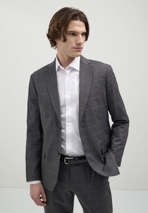 Giovane uomo che indossa un completo grigio a quadri e una camicia bianca, in piedi con una mano in tasca, mentre guarda di lato su uno sfondo semplice.