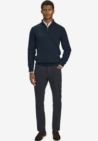 Marinblå kabelstickad tröja med dragkedja, matchad med mörkgrå byxor. Outfitten har en smal passform och en kontrasterande vit skjorta under.