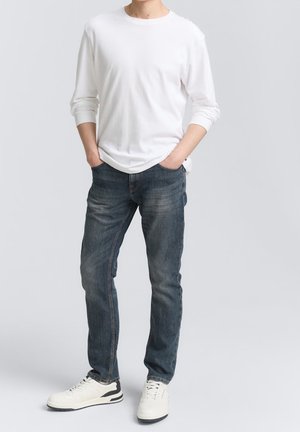 Homme portant une chemise blanche à manches longues, un jean bleu et des baskets blanches, debout les mains dans les poches devant un fond uni.