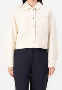 Chaqueta de denim color crema, cortada, con cuello, dos bolsillos en el pecho, botones de presión plateados y una textura sutil. Lleva puesta con pantalones oscuros.