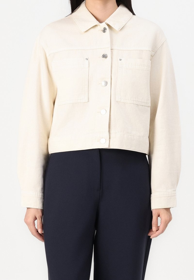 Chaqueta de denim color crema, cortada, con cuello, dos bolsillos en el pecho, botones de presión plateados y una textura sutil. Lleva puesta con pantalones oscuros.