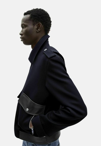 Giacca in misto lana blu navy con tasca anteriore in pelle sintetica, polsini con zip e colletto strutturato. La vista laterale mette in risalto la forma su misura e la texture.