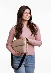 Béžová crossbody taška s hladkým povrchem a sklopenou klopou, vybavená černým nastavitelným popruhem. Model má na sobě růžový žebrovaný svetr a modré džíny.