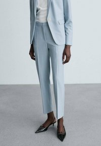 Abito su misura di un blu pallido con pantaloni slim-fit e giacca a un bottone, abbinato a tacchi neri a punta. Tessuto liscio.