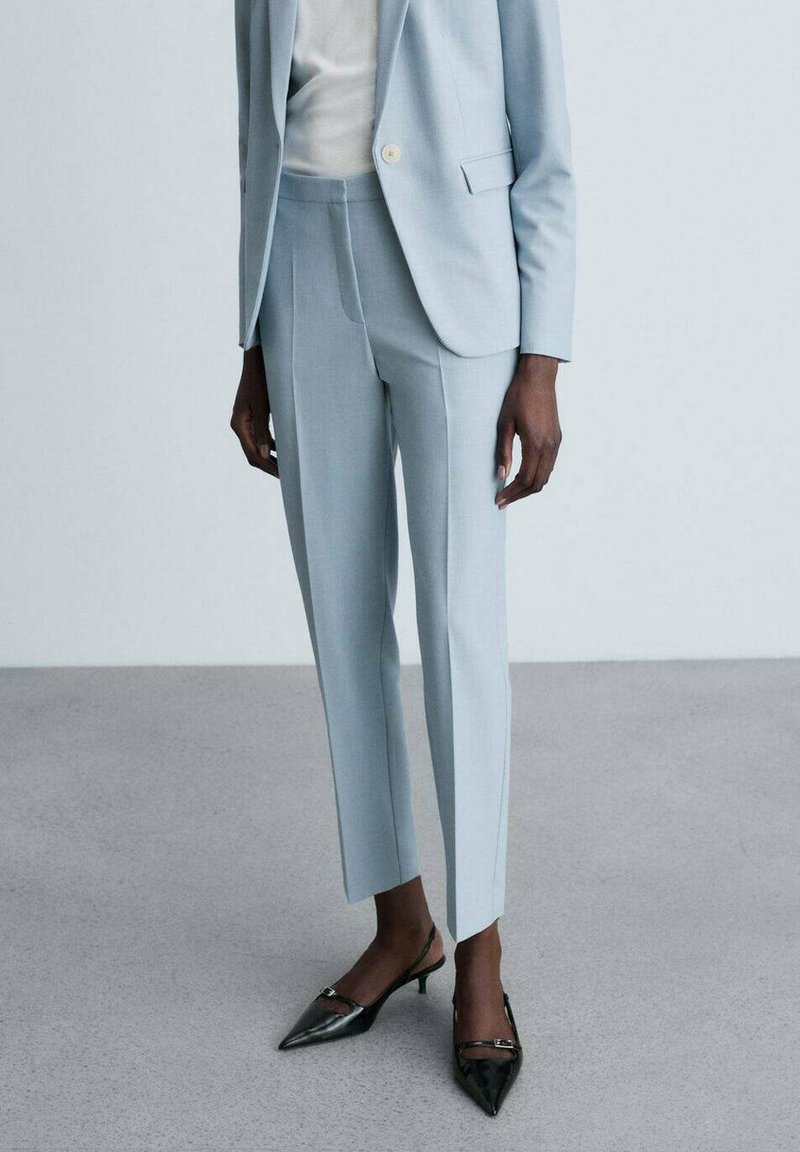 Abito su misura di un blu pallido con pantaloni slim-fit e giacca a un bottone, abbinato a tacchi neri a punta. Tessuto liscio.