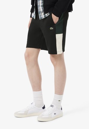 Pantalones cortos negros con acentos laterales verdes y crema, que presentan un pequeño logo de Lacoste. Combinados con zapatillas blancas y calcetines hasta los tobillos.