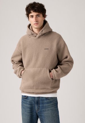 Beige fleece pullover hoodie with drawstring hood, front pocket, and small "Levi's" logo. Worn over a white shirt and blue jeans.