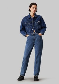 Chaqueta de mezclilla azul con un diseño recortado, contrastada con jeans azules de cintura alta. Presenta costuras prominentes y dos bolsillos en el pecho.
