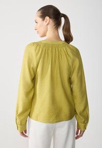 Gul langermet bluse med teksturert overflate, samlet hals og knappede mansjetter. Bakdelen viser en subtil rutemønstring.