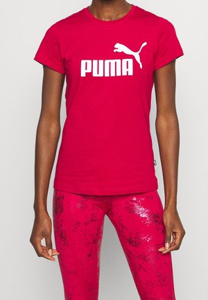 Person trägt ein rotes Puma T-Shirt mit weißem Logo und passende rote gemusterte Leggings, steht vor einem schlichten Hintergrund.