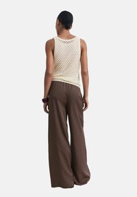 Top sin mangas de malla color crema con un diseño de punto abierto, combinado con pantalones anchos de pierna suelta en color marrón. El conjunto presenta una silueta relajada y una textura sutil.
