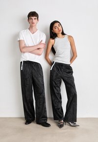 Unisex black pants with a shiny texture, featuring a drawstring waist and side pockets. Grey sleeveless top and white v-neck t-shirt are worn.