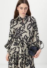 Abito a maniche lunghe con motivo nero e beige a foglie, con una cintura in vita. La modella tiene in mano una piccola borsa nera arricciata.