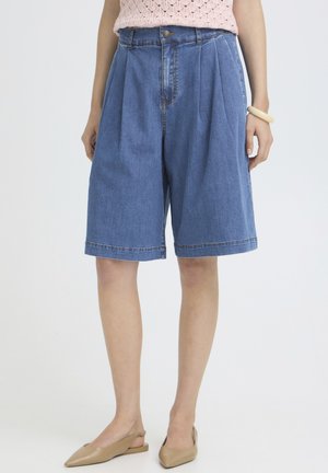 Kvinde iført højtaljede blå denim knælange shorts med plisseringer, en lyserød strikket top, beige slingback-flats og et cremefarvet armbånd.