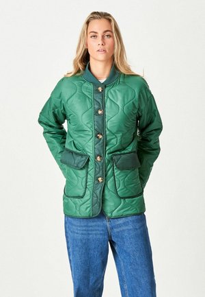 Tânără purtând o jachetă verde matlasată cu nasturi și buzunare cu clapete, alături de blugi albaștri, stând pe un fundal simplu, deschis la culoare.