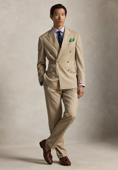 Uomo in piedi con le gambe incrociate, indossa un abito beige doppiopetto, camicia blu, cravatta a righe, fazzoletto verde nel taschino e mocassini marroni con nappine.