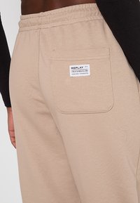 Sandfarbene Jogginghose aus weichem Material, mit elastischem Bund und einer Gesäßtasche, auf der ein weißes Etikett mit dem Markennamen aufgedruckt ist.