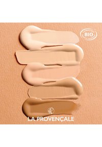 Cinq échantillons de fond de teint dans des tons beige variés disposés horizontalement, sur un fond texturé en pêche. Comprend un logo circulaire et le nom de la marque.