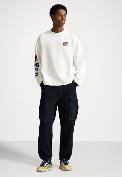 Sweatshirt branca com um pequeno logótipo de bandeira no peito e texto azul "USA" numa das mangas, combinada com calças cargo pretas e ténis coloridos.