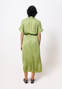 Satin midi-Kleid in Grün mit kurzen Drop-Ärmeln, tailliertem Gürtel und glatter Textur. Es hat einen geraden Saum und wird mit gemusterten flachen Schuhen getragen.