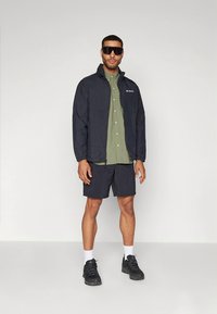 Chaqueta impermeable azul marino sobre una camisa de botones color oliva, combinada con pantalones cortos oscuros y zapatillas negras, con gafas de sol reflectantes.