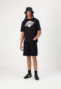Svart T-skjorte med hvitt grafisk trykk og svarte shorts. Brukt med svarte joggesko med hvite striper og en svart bøttehatt.