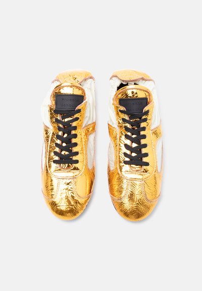 Ζευγάρι λαμπερών χρυσών και λευκών αθλητικών παπουτσιών με μαύρα κορδόνια, φωτογραφημένα από πάνω σε απλό φωτεινό φόντο.