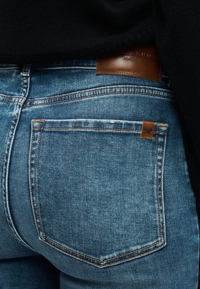 Κοντινό πλάνο πίσω τσέπης μπλε τζιν με καφέ δερμάτινη ετικέτα μάρκας και μικρό ραμμένο ταμπελάκι με πεταλούδα.