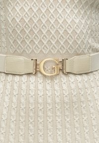 Ζώνη σε ανοιχτό κρεμ χρώμα, με ελαστική υφασμάτινη λωρίδα. Διακριτή αγκράφα με χρυσό λογότυπο "G", ομαλή υφή και διακριτικό σχεδιασμό με μοτίβα.