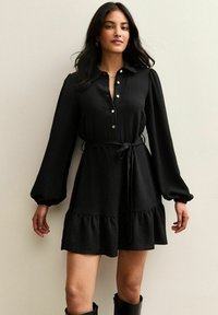 Vestido negro de botones con mangas largas abullonadas, cintura ajustable con lazo y un dobladillo con volantes. Fabricado con una tela texturizada y detalles en botones en tono dorado.