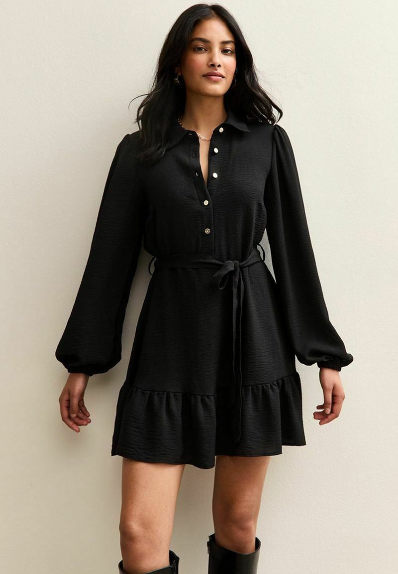 Vestido negro de botones con mangas largas abullonadas, cintura ajustable con lazo y un dobladillo con volantes. Fabricado con una tela texturizada y detalles en botones en tono dorado.