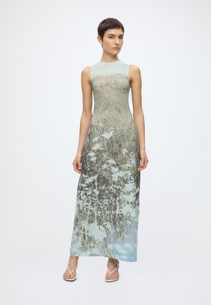 Femme debout portant une robe longue sans manches avec un imprimé inspiré par la nature représentant de l'herbe et des fleurs, et des sandales à fines brides.