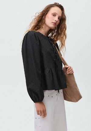 Blusa nera con texture, maniche a sbuffo e orlo arricciato. Presenta un laccio al collo e decorazioni sottili. Pantaloni bianchi semplici e una borsa beige.