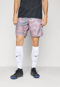 Mannlig idrettsutøver iført grå og røde mønstrede shorts, hvite Nike leggskinn, svarte fotballsko med blå stifter, og en svart sportsskjorte.