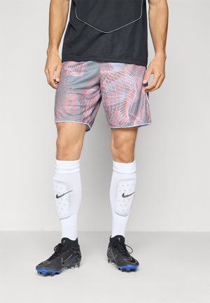 Männlicher Athlet trägt graue und rote gemusterte Shorts, weiße Nike Schienbeinschoner, schwarze Fußballschuhe mit blauen Stollen und ein schwarzes Sportshirt.
