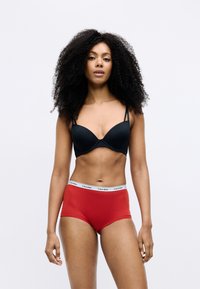 Fekete melltartó formázott kosarakkal és állítható pántokkal, piros alsóneművel, melyen egy fehér Calvin Klein logós derékpánt található. Sima textúra.